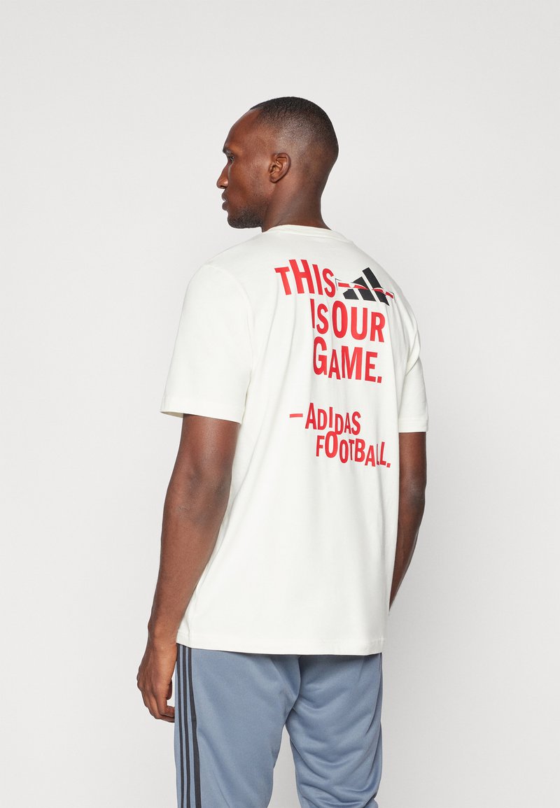 Hvid kortærmet T-shirt med rød tekst "DETTE ER VORES SPIL. - Adidas Fodbold." på ryggen, med rund hals og en afslappet pasform.