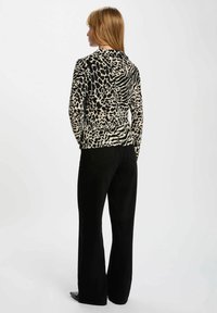Donna con capelli biondi lisci che indossa una maglietta a maniche lunghe con stampa animale nera e beige e pantaloni larghi neri, in piedi di spalle.