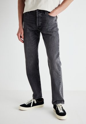 Person trägt dunkelgraue Jeans mit geradem Schnitt und schwarze flache Turnschuhe mit weißen Schnürsenkeln und Sohlen, steht mit einer Hand in der Tasche.