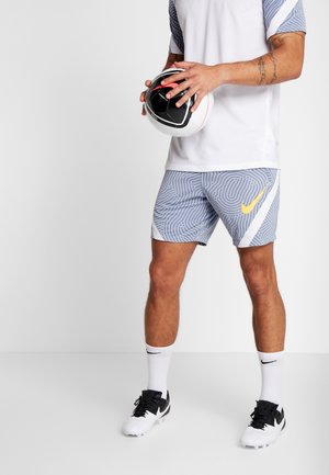 Mann in gemusterten grauen Sportshorts und weißem Hemd hält einen schwarz-weißen Fußball vor einfachem Hintergrund.