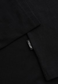 Tissu noir plié avec une petite étiquette de vêtement noire portant le texte "LEVEL UP" en blanc cousue dans la couture.
