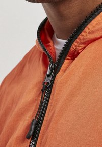 Orange jakke med en tekstureret overflade, der har en sort lynlås og krave. Stoffet ser let og en smule glinsende ud.