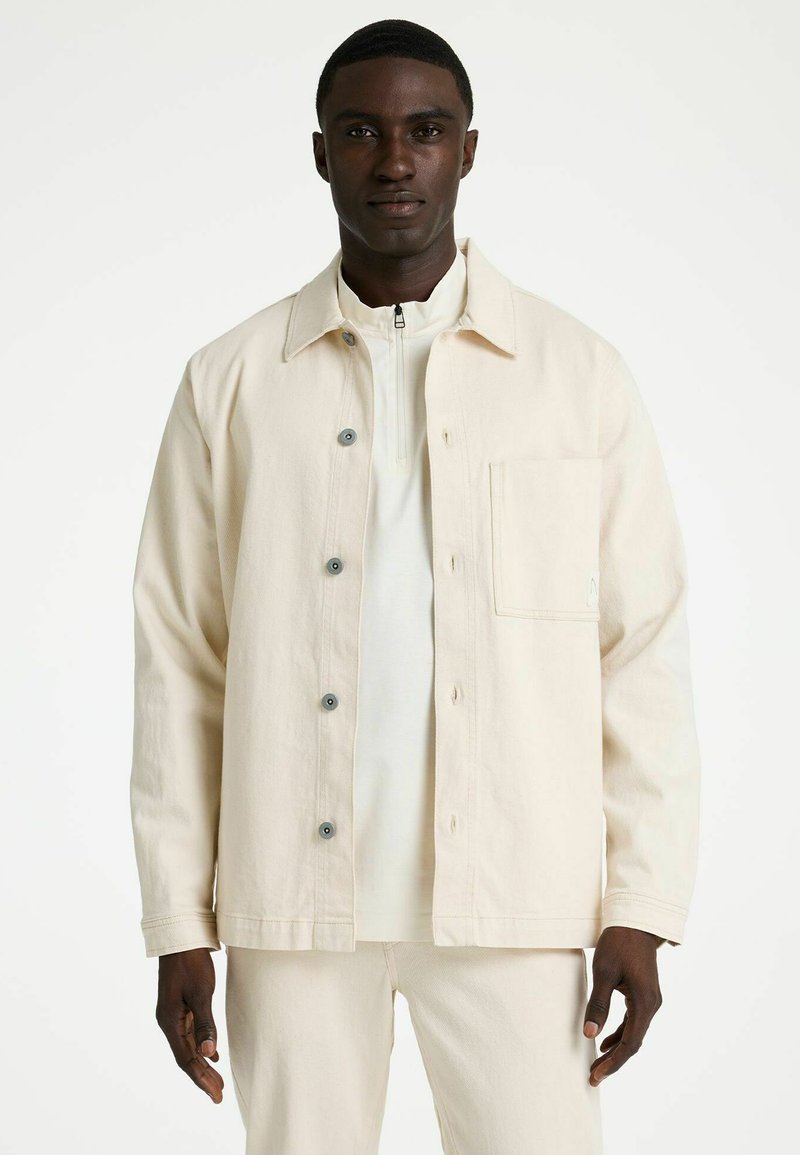 Kreemika värvi jakk pehmest kangast, kraega kaelusega, ees nööbid ja rinnatasku. Modell kannab all õhukest valget toppi.