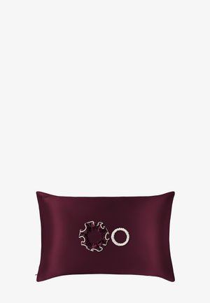 Cojín rectangular rojo oscuro con una scrunchie con volantes y una pulsera circular blanca con cuentas colocada encima.