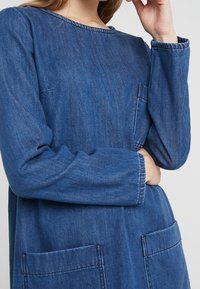 Tummansininen farkkutunika, jossa on pyöreä pääntie, pitkät hihat ja kaksi etutaskua. Ompeleet korostavat saumoja.