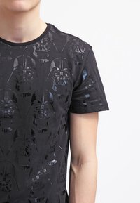 Černé bavlněné tričko s raženým vzorem hlav Dartha Vadera, krátké rukávy a kulatý výstřih. Měkká textura s lesklým povrchem.