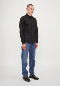 Vyras stovi apsirengęs juoda mygtukine marškinėliais, mėlynomis džinsais ir juodais kvadratiniais batais ant paprastos baltos fono.