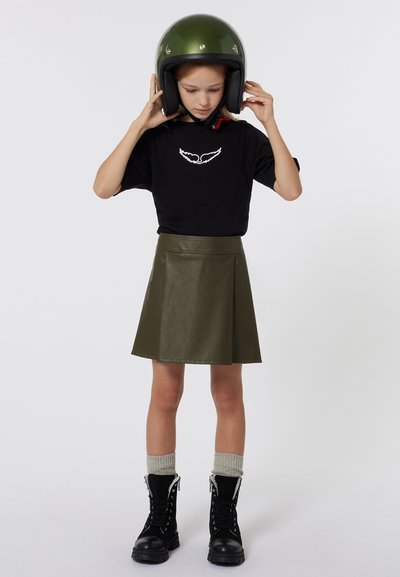 T-shirt noir avec broderie d'ailes blanches, jupe en simili cuir vert olive, chaussettes grises et bottes noires à lacets avec accents en argent, surmonté d'un casque vert.