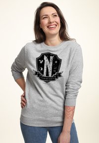 Mujer sonriente con cabello largo castaño lleva una sudadera gris con un logo de escudo negro y texto en latín, de pie con la mano en la cadera frente a un fondo claro.