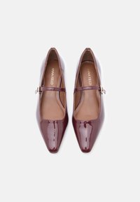 Μπορντό παπούτσια Mary Jane από πατενταρισμένο δέρμα με μυτερή μύτη, λουράκι στον αστράγαλο και λεπτομέρεια από χρυσό τόνο στη θηλιά. Λείος υφή και κομψός σχεδιασμός.