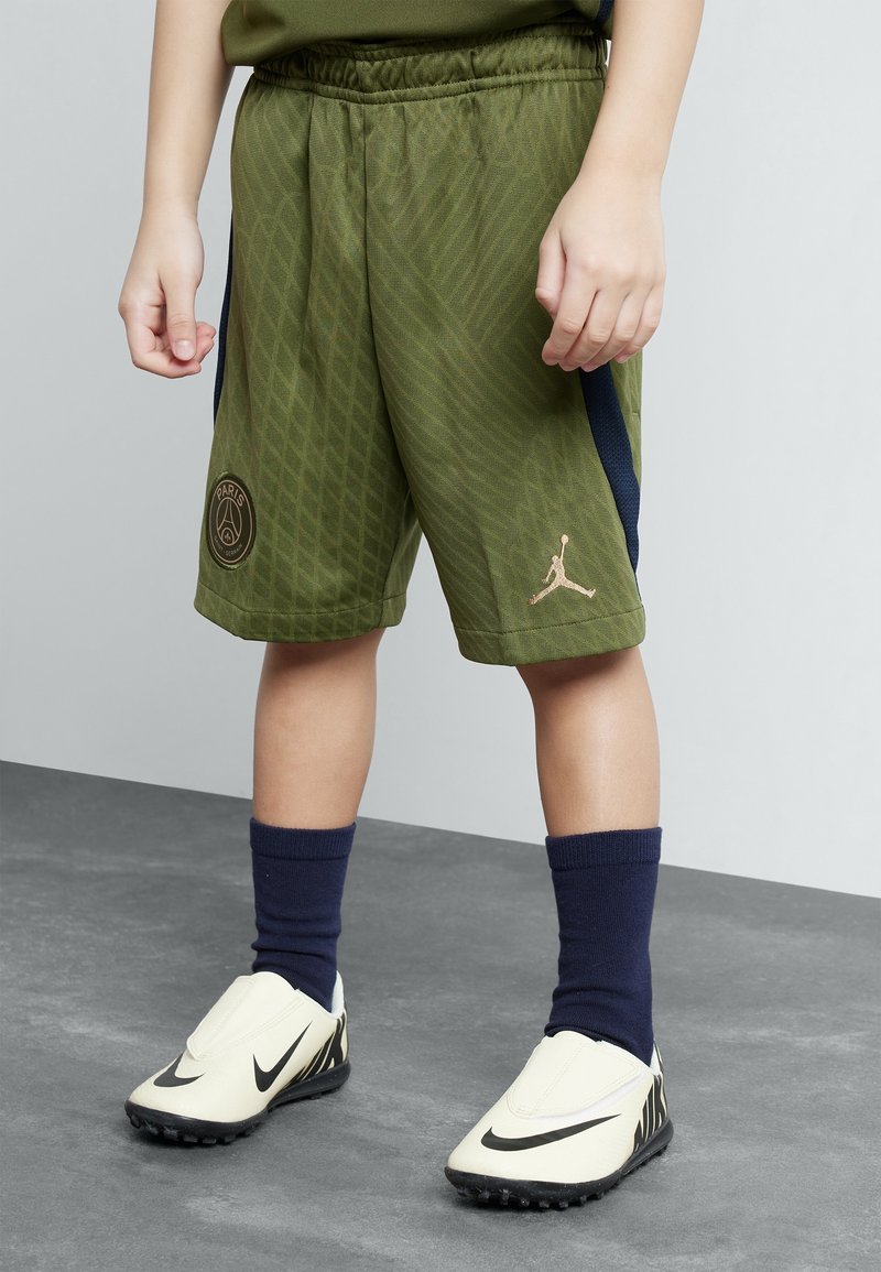 Lapsi, joka pukeutunut oliivinvihreisiin urheilushortseihin, joissa on Paris- ja Jordan-logot, tummansinisiin sukkiin ja valkoisiin Nike-nappulakenkiin, seisoo harmaalla lattialla.