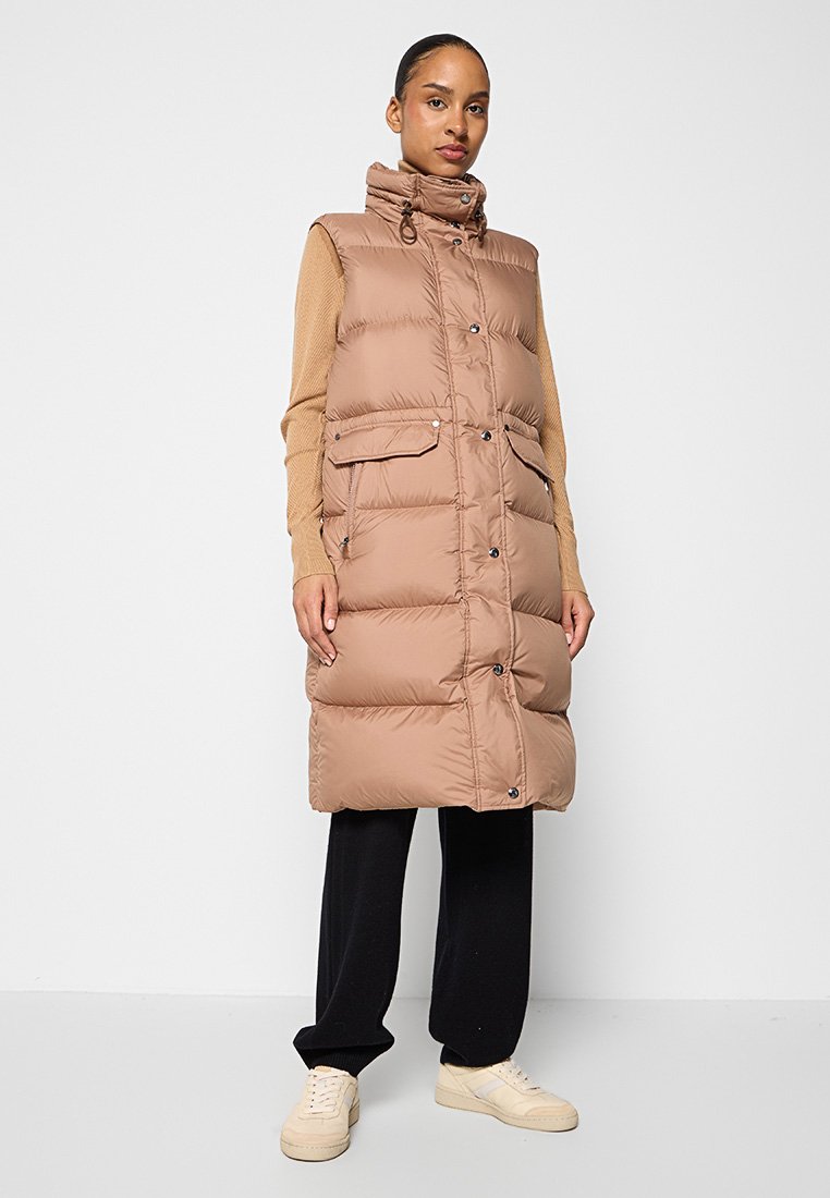 Braune ärmellose Steppweste mit hohem Kragen, vorderen Knöpfen und zwei seitlichen Taschen. Kombiniert mit einem beigen Pullover und schwarzen Hosen.