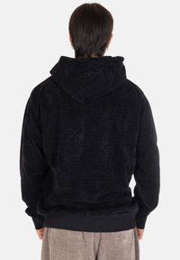 Felpa in corduroy nera con un cappuccio spazioso, polsini a coste e una superficie texturizzata, indossata con pantaloni di colore chiaro, vista da dietro.