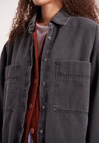 Osebo ose kodraste lase, oblečen v več slojev oblačil: belo srajco, rjavo-rjavo pleteno jopico z gumbi in preveliko temno sivo denim jakno z žepi.