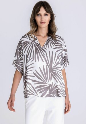 Vrouw draagt een losse blouse met een wit en grijs palmbladpatroon, een V-hals en korte mouwen, gecombineerd met een witte broek tegen een effen achtergrond.
