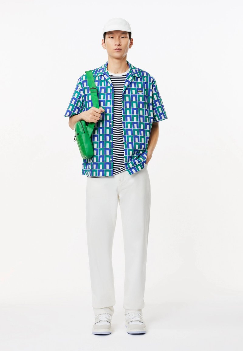 Chemise à manches courtes avec un motif géométrique bleu et vert, tee-shirt en noir et blanc à rayures en dessous, pantalon blanc et sac bandoulière vert.