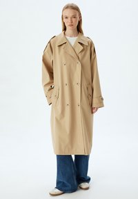 Beige dubbelknäppt trenchcoat med stor krage, knappdetaljer och sidofickor. Stylad med vida blå jeans och vita sneakers.