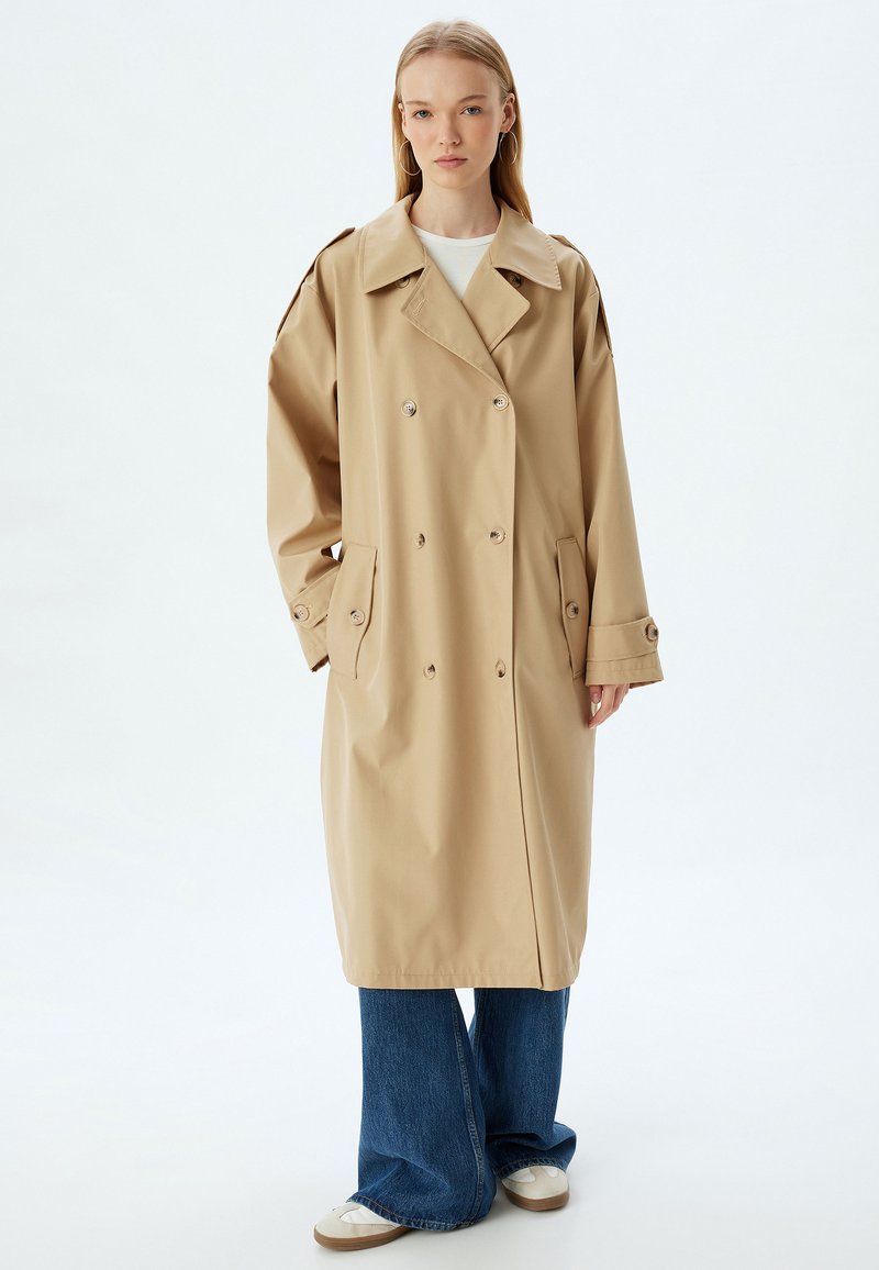 Beige dubbelknäppt trenchcoat med stor krage, knappdetaljer och sidofickor. Stylad med vida blå jeans och vita sneakers.