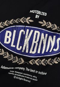 Zwarte stof met een circulair blauw embleem met verweerd witte tekst "BLCKBNNS," omringd door een beige laurierontwerp en aanvullende tekst eronder.