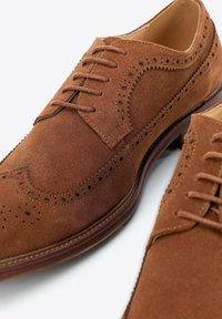 Zapatos brogue de piel de ante marrón con detalles perforados y cordones, mostrados en primer plano sobre un fondo blanco.
