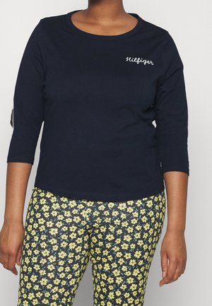 Personne portant une chemise bleu marine à manches trois-quarts avec le texte « Hilfiger » et un pantalon noir avec de petits motifs de fleurs jaunes.