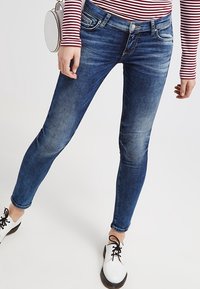 Jeans de denim azul ajustados con ligero desgastado, diseño estándar de cinco bolsillos y presillas para cinturón, combinados con zapatos blancos y una camiseta a rayas.