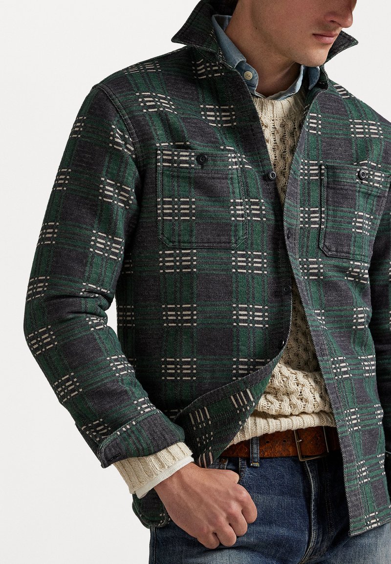 Mand iført grøn og grå ternet skjorte med knapper over en lys kabelstrik sweater og blå jeans med brun bælte.