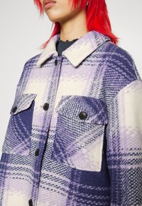 Person mit roten Haaren, der eine lila-weiße karierte Knopfjacke mit großen Taschen und schwarzen Knöpfen trägt.