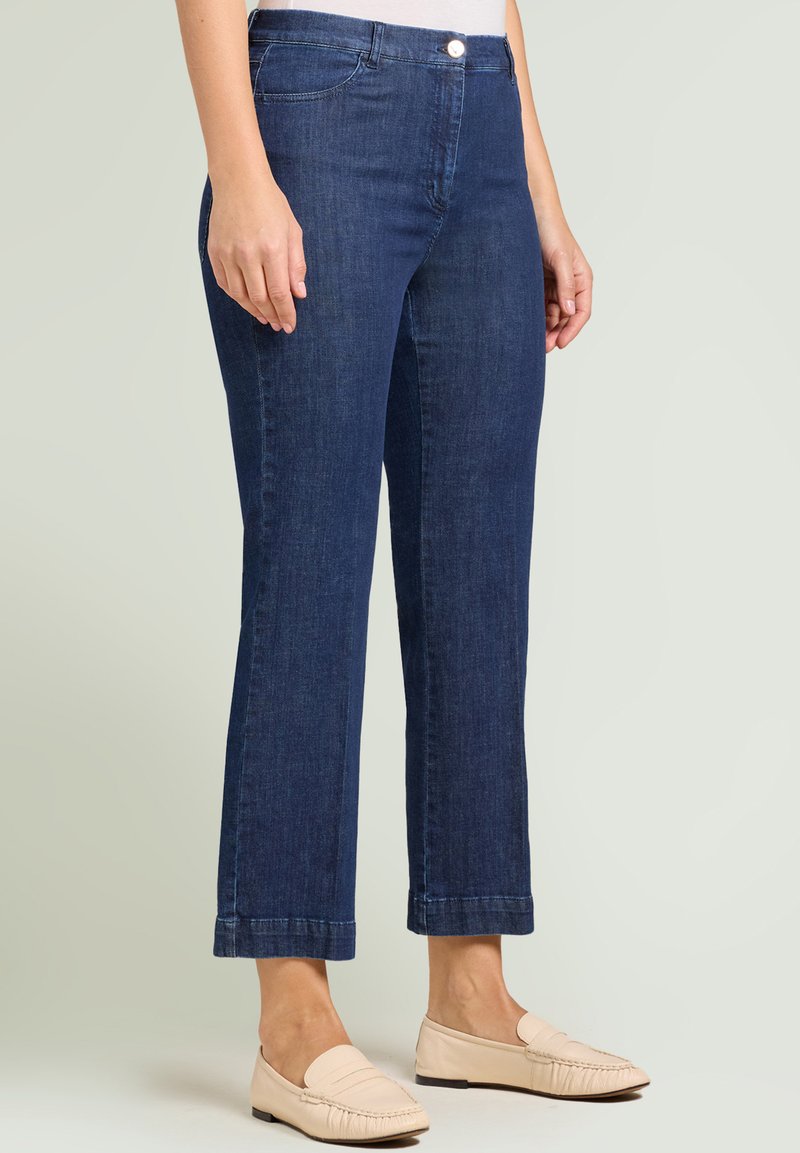 Nainen, jolla on suoralahkeiset tummansiniset farkut käännetyillä lahkeilla ja beiget slip-on-mokkasiinit, seisoo vaaleaa taustaa vasten.