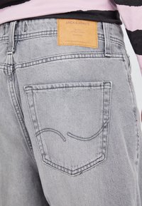 Jeans de mezclilla gris con un bolsillo trasero que presenta un detalle de costura curva y una etiqueta de cuero marrón con la inscripción "JACK & JONES".