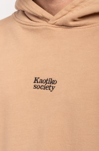 Hanorac bej dintr-un material moale, având un logo brodat negru "Kaotiko society" pe partea stângă a pieptului.