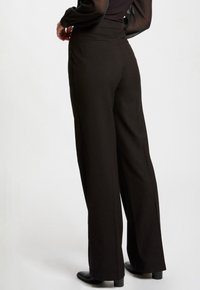 Pantalons larges noirs avec une texture lisse, taille mi-haute et deux poches arrière ; associés à des bottes à talons jusqu'à la cheville.