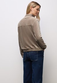 Giacca zip-up in velluto taupe con colletto alto, vita elastica e dettagli di cucitura discreti, abbinata a jeans blu scuro.