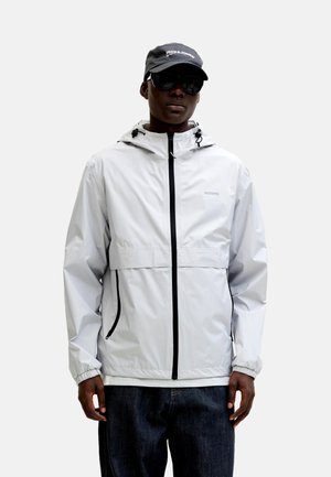 Personne portant une veste à capuche zippée gris clair avec des fermetures éclair noires, des lunettes de soleil foncées, une casquette noire avec un logo blanc, et un pantalon foncé sur fond blanc.