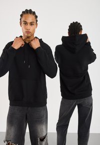 Svart hettegenser laget av myk stoff, med en frontlomme, snor i hetten og avslappet passform, parret med slitte grå jeans.