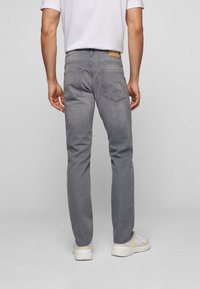 Man som bär grå jeans, vita sneakers och en vit T-shirt som står vänd bort mot en neutral grå bakgrund.