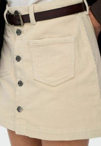 Jupe en velours côtelé beige avec une fermeture à cinq boutons sur le devant, deux poches cousues et une texture tissée. Ceinturée avec une ceinture en cuir marron foncé.