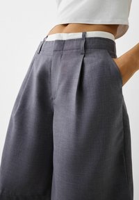 Šedé plisované kraťasy s texturovanou tkaninou, bílým pasem a bočními kapsami, s uvolněným střihem a přizpůsobeným designem.