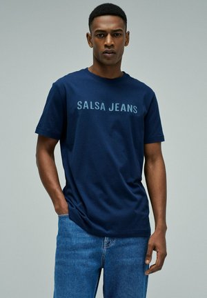 Hombre vistiendo camiseta azul marino con el logo "SALSA JEANS" y vaqueros azules, de pie con una mano en el bolsillo contra un fondo gris liso.