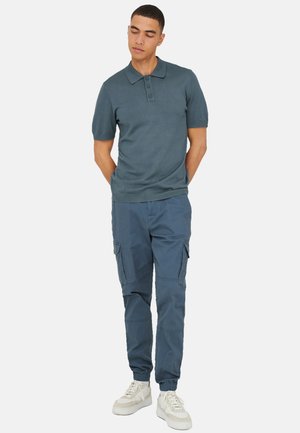 Man staat met handen achter de rug, draagt een blauw poloshirt met korte mouwen, blauwe cargobroek en witte sneakers op een witte achtergrond.