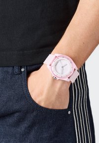 Okrogla ura s rožnato silikonsko narukvico in belo številčnico; ima minimalne oznake in kovinsko krono, nosi se na zapestju blizu jeansa.