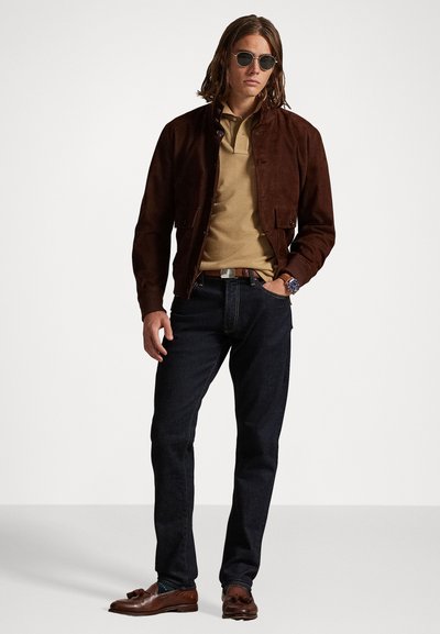 Καφέ σουέτ jacket, κοντός γιακάς, κούμπωμα με κουμπιά, σε συνδυασμό με μπεζ πουκάμισο, σκούρα τζιν και καφέ λαστιχένια παπούτσια. Φορετά κυκλικά γυαλιά ηλίου.