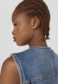 Gilet di jeans con una tonalità blu, design senza maniche e dettagli di cucitura visibili. Capelli acconciati in trecce strette, decorati con piccoli orecchini.