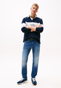 Noor mees seisab käed rüpes, seljas tumesinine-valge pullover, sinised teksad ja sinised tossud valgel taustal.