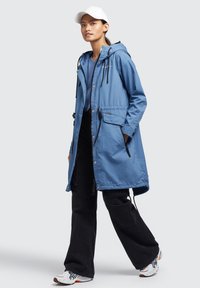 Parka ușor de culoare albastră, cu glugă, șnururi ajustabile, buzunare laterale și închidere cu capse, asortată cu pantaloni negri largi și adidași.