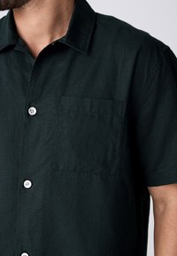 Camisa de manga corta verde oscuro en mezcla de algodón, que presenta un cierre delantero con botones, botones blancos y un único bolsillo en el pecho.