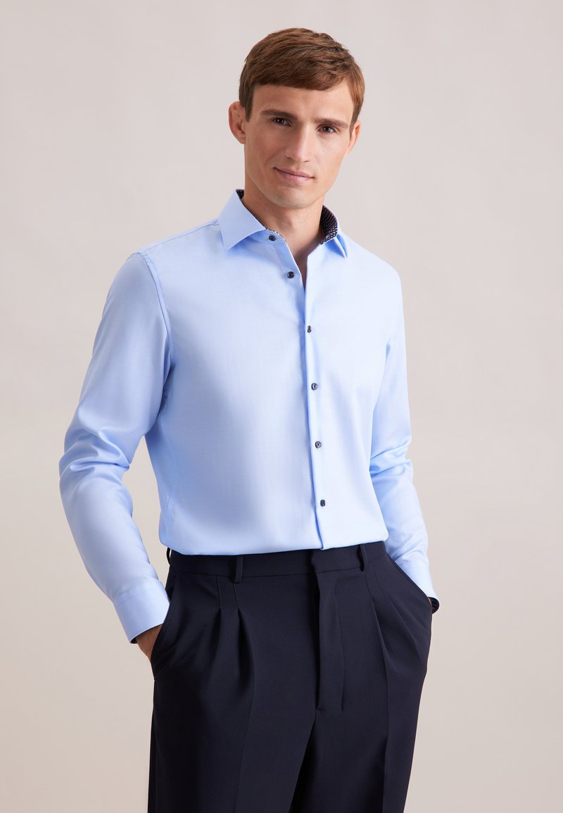 Camicia azzurra chiara con colletto aperto e bottoni scuri. La camicia ha maniche lunghe, con un dettaglio a fantasia contrastante all'interno del colletto.