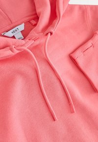 Koralno roza jopa s kapuco z vrvico, mehka tkanina in rebrast manšet. V notranjosti je etiketa z znamko 'NEXT'.