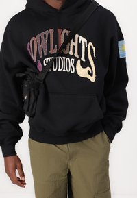 Schwarzer Kapuzenpullover mit besticktem "OWLIGHTS STUDIOS" Schriftzug, Kängurutasche, kombiniert mit olivgrünen Cargohosen und einer kleinen Umhängetasche.