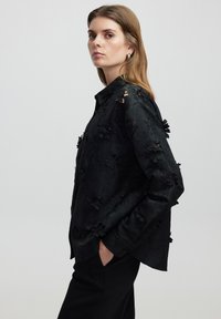 Zwarte langemouwen shirt met bloemvormige uitsparingen, een gladde textuur en een relaxte pasvorm. Knopsluiting en zijzakken zichtbaar.